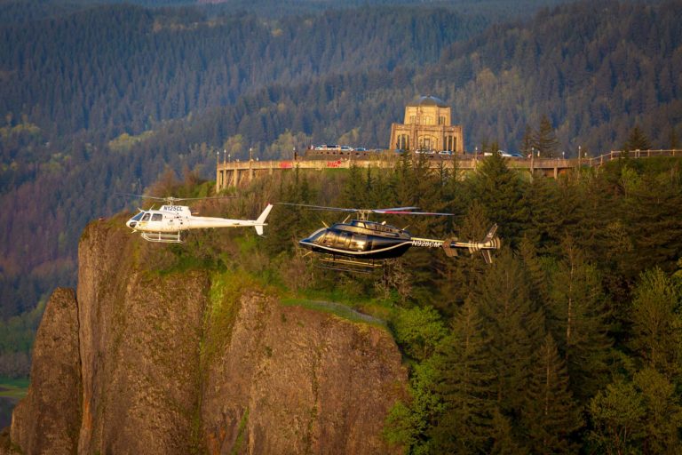 two helicopters flying across forested landscape and cliffside building